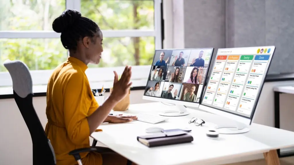 Mujer frente a 2 pantallas, una con una reunión de zoom abierta, la otra con un kanban board a la vista